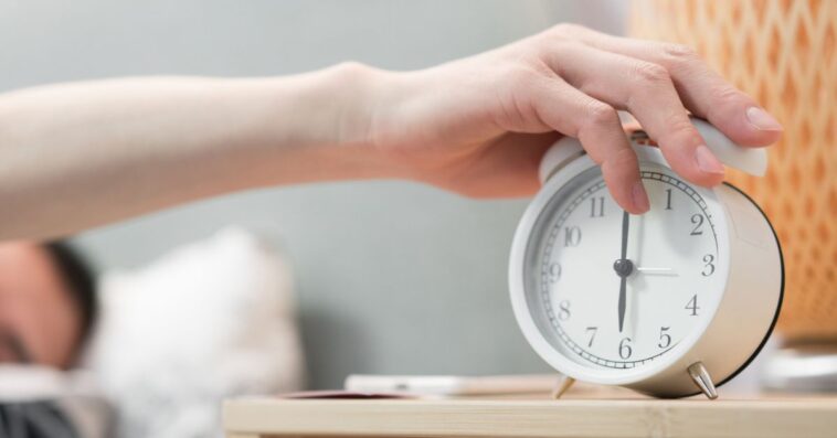 An asleep man is turning off alarm clock.
