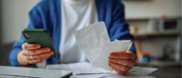 Unrecognizable woman with a smartphone, laptop, and checks at a table at home.