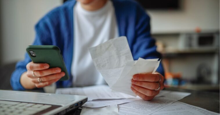 Unrecognizable woman with a smartphone, laptop, and checks at a table at home.