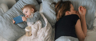 A woman lying down in bed next to a baby holding a smartphone.