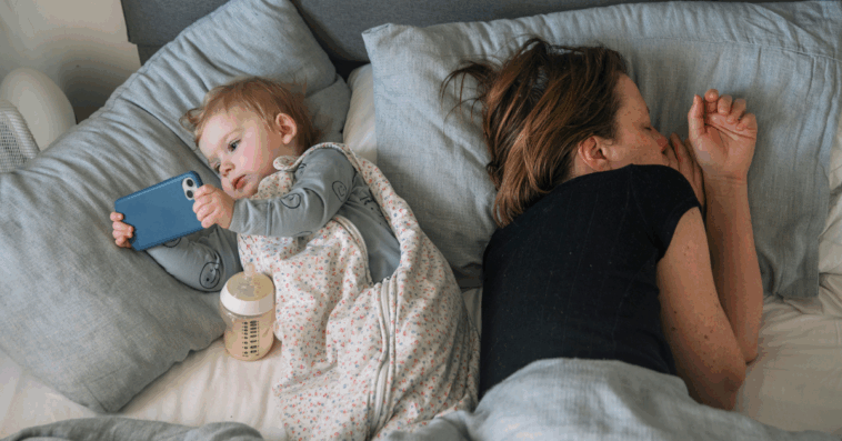 A woman lying down in bed next to a baby holding a smartphone.