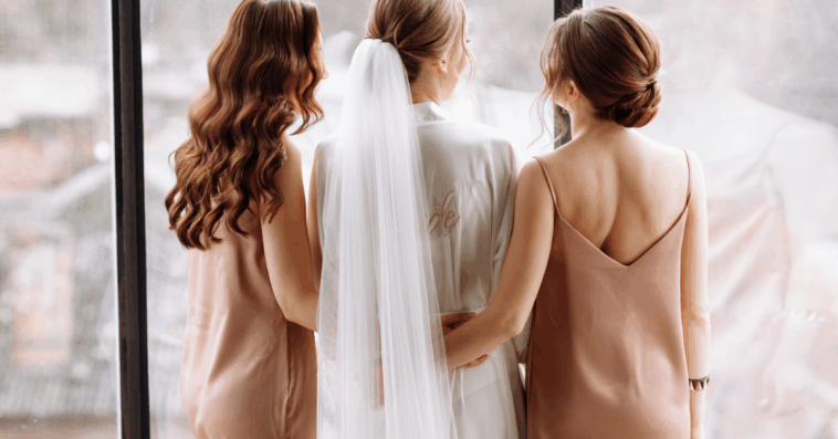 A woman in a wedding veil surrounded by two women in tan silk dresses.