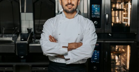 Portrait of confident, young male chef wearing a white uniform and standing with his arms crossed in the kitchen of a restaurant.