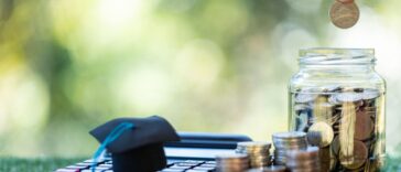 A calculator, stacks of coins and coin jar sit on the grass. Two fingers drop a coin into the jar.
