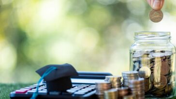 A calculator, stacks of coins and coin jar sit on the grass. Two fingers drop a coin into the jar.