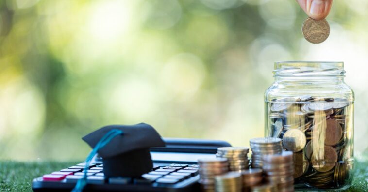 A calculator, stacks of coins and coin jar sit on the grass. Two fingers drop a coin into the jar.