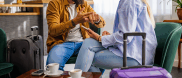 A couple sitting at a table talking to each other at a table with cups of coffee on it and luggage in front of them.