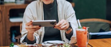 Smiling young woman taking photos of her scrumptious and beautifully plated dishes on the table with her smartphone in outdoor cafe. Enjoying her meal and sharing foodie photos on social media. Digital dining. People, food, lifestyle and technology.