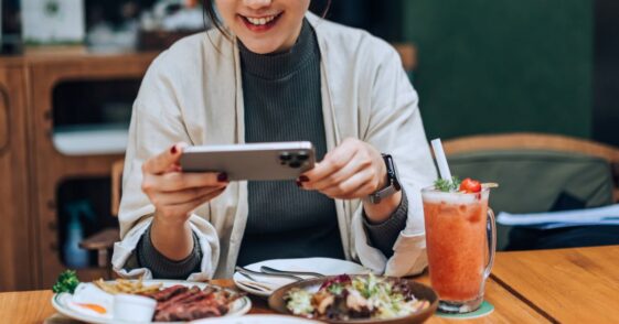 Smiling young woman taking photos of her scrumptious and beautifully plated dishes on the table with her smartphone in outdoor cafe. Enjoying her meal and sharing foodie photos on social media. Digital dining. People, food, lifestyle and technology.