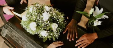 High angle shot of unrecognizable hands on black wooden coffin with white roses on top, pastor holding holy book.
