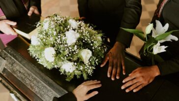 High angle shot of unrecognizable hands on black wooden coffin with white roses on top, pastor holding holy book.