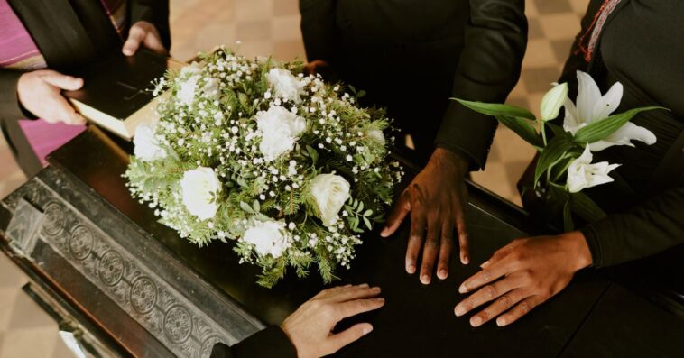 High angle shot of unrecognizable hands on black wooden coffin with white roses on top, pastor holding holy book.