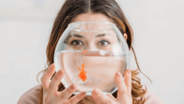 A woman holding a fishbowl in front of her face with a swimming goldfish in it.