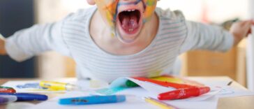 A boy paints his face with colorful felt pens, smiles and stares into the camera.