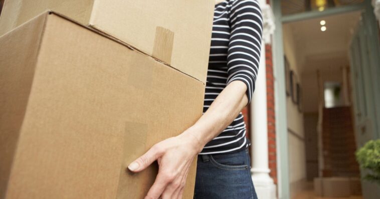 Unidentifiable woman carrying brown boxes from house.
