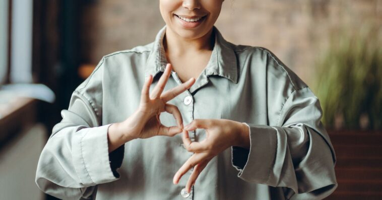 Smiling young woman using sign language, demonstrating the letter r with her hands while standing in a stylish modern loft apartment filled with natural light.