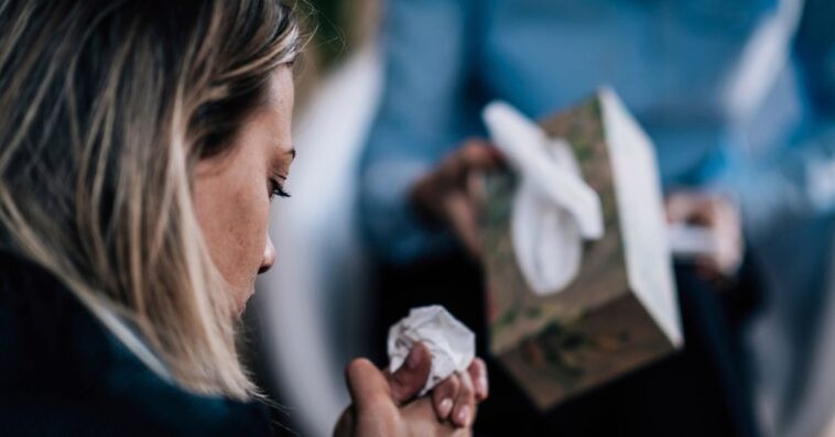 Back shot of a troubled woman crying, talking about her problems. A person in the background offers her tissues.