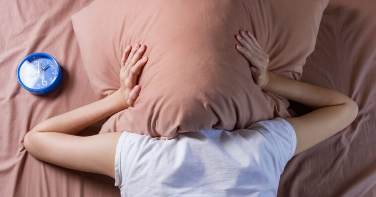 A woman covers her head with a pillow. An alarm clock sits next to her.