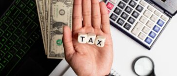 Businessman making the word tax on wooden blocks on an office table with laptop, calculator, US dollars and diary on it.