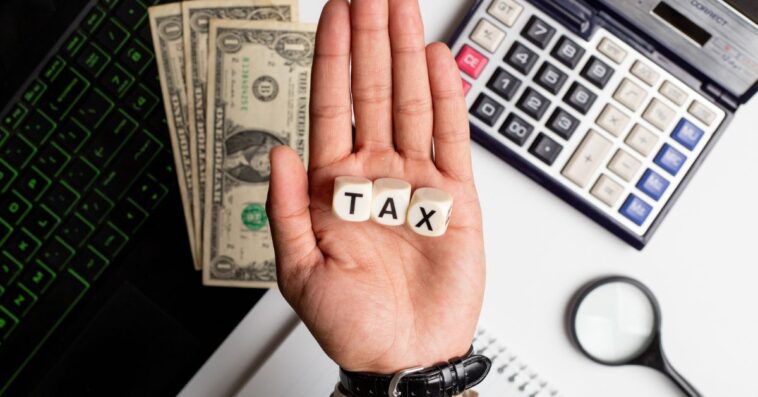 Businessman making the word tax on wooden blocks on an office table with laptop, calculator, US dollars and diary on it.