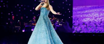 A shot of Taylor Swift singing during The Eras Tour. She wears a floor length, sparkling light blue dress.