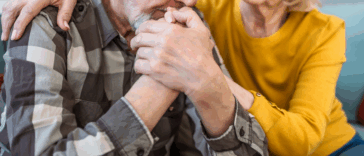 An older man resting his head on his hands and a woman leaning in to comfort him.