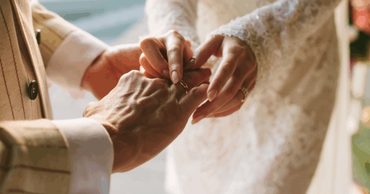 A groom sliding a ring on the finger of a bride.