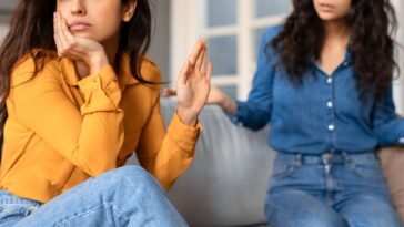 Offended young woman in a yellow top appears upset while conversing with another lady friend in a blue shirt.