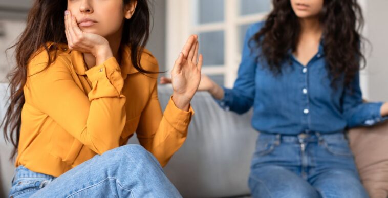 Offended young woman in a yellow top appears upset while conversing with another lady friend in a blue shirt.
