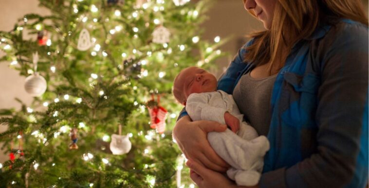 Mother carrying newborn baby while standing in front of a Christmas tree at home.