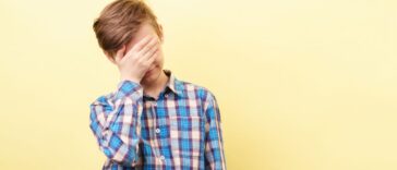 Facepalm, desperate boy standing in front of a yellow background, covers his face with his hand.