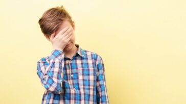 Facepalm, desperate boy standing in front of a yellow background, covers his face with his hand.