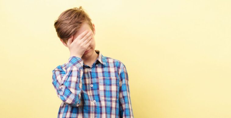 Facepalm, desperate boy standing in front of a yellow background, covers his face with his hand.