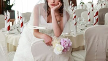 A sad bride sits in an empty banquet room prepared for a wedding, she holds flowers.