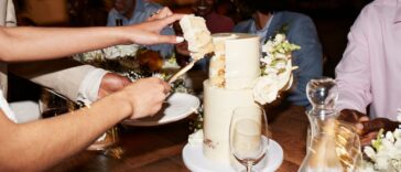 Cropped hands of newlywed couple cutting wedding cake near guests sitting at illuminated dining table during reception dinner party.