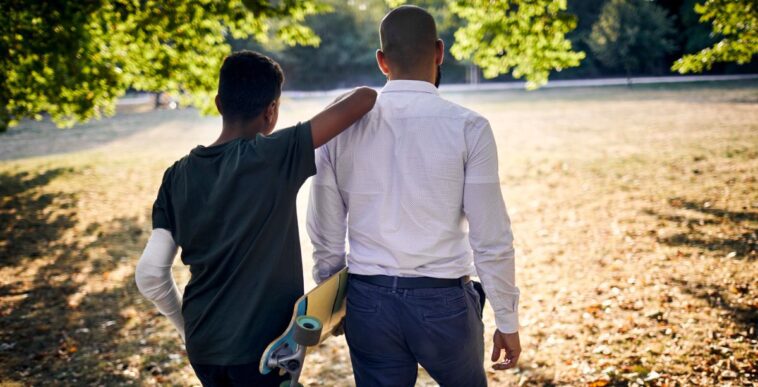 Rear view of a father and son with skateboard walking in a park.