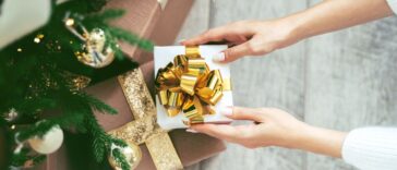 Tender woman hands putting white wrapped gift box with golden bow tie under the Christmas tree to other presents.