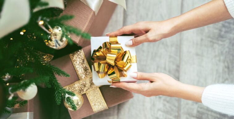 Tender woman hands putting white wrapped gift box with golden bow tie under the Christmas tree to other presents.