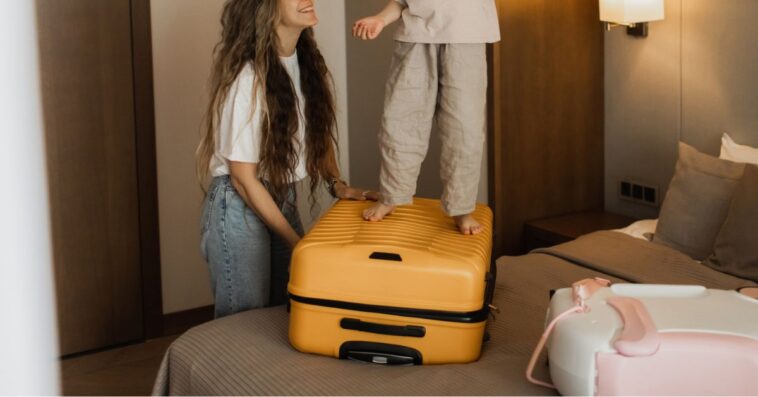 Kid is messing around in hotel room, jumping on bed, while mother is unpacking luggage.