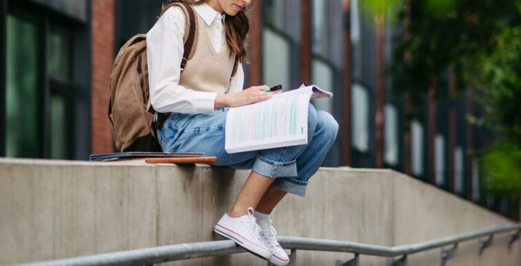Portrait of female student sitting on low wall in front of university building, studying, reading textbook, preparing for final exam.