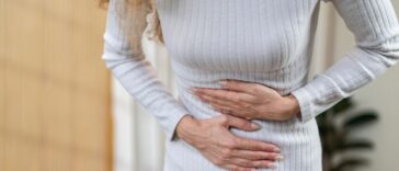 Close-up of a woman's midsection. She is wearing a white dress and is holding her stomach in pain.