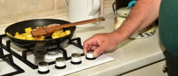 A man turns the knob of a gas stove to turn off the heat on the burner underneath a frying pan.