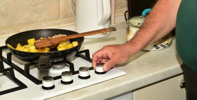 A man turns the knob of a gas stove to turn off the heat on the burner underneath a frying pan.