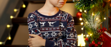 A child with a frown on his face, folding his arms, standing in front of a Christmas tree.
