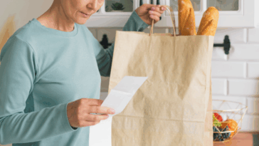 A woman at a counter looking at a list next to a bag full of groceries.