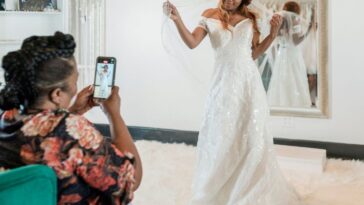A woman posing while wearing a wedding dress and veil during a fitting in a bridal store, with her proud mother taking photos using her smartphone.