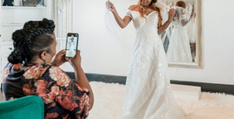 A woman posing while wearing a wedding dress and veil during a fitting in a bridal store, with her proud mother taking photos using her smartphone.