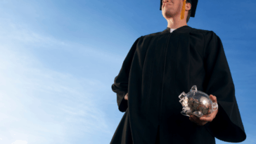 A graduate holding a piggy bank looking off into the sky.