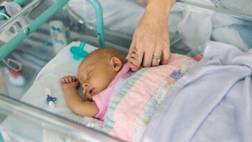 A young baby girl lies in a hospital cot. She is suffering from neonatal jaundice. Her mother holds her hand and comforts her as she sleeps.