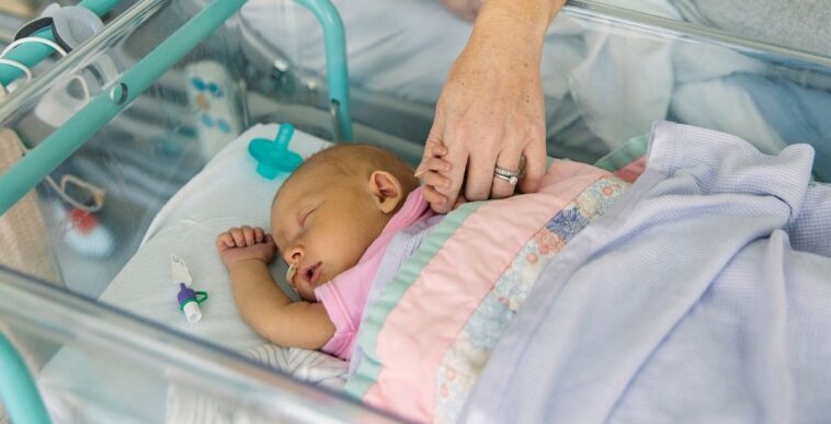 A young baby girl lies in a hospital cot. She is suffering from neonatal jaundice. Her mother holds her hand and comforts her as she sleeps.